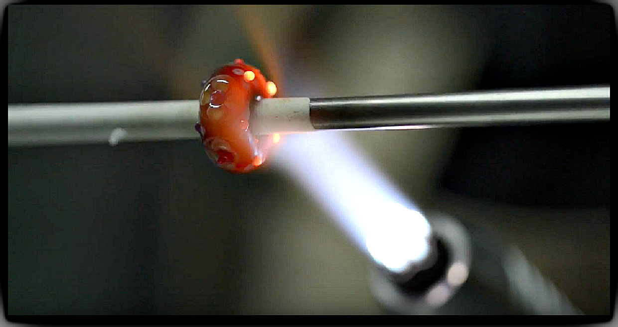 A hot flame with melted glass on a metal rod to show how glass beads are made.