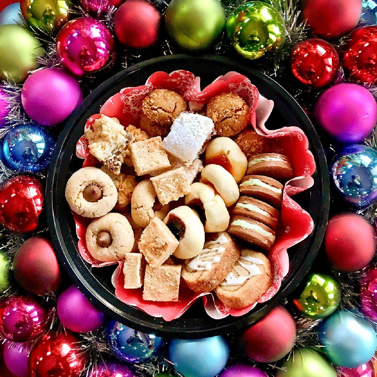 A container of Christmas cookies with bright coloured baubles around it.