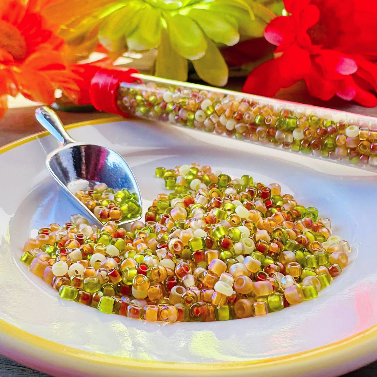 A mix of colourful seed beads, loose on a white plate and in a plastic tube, and flowers in background