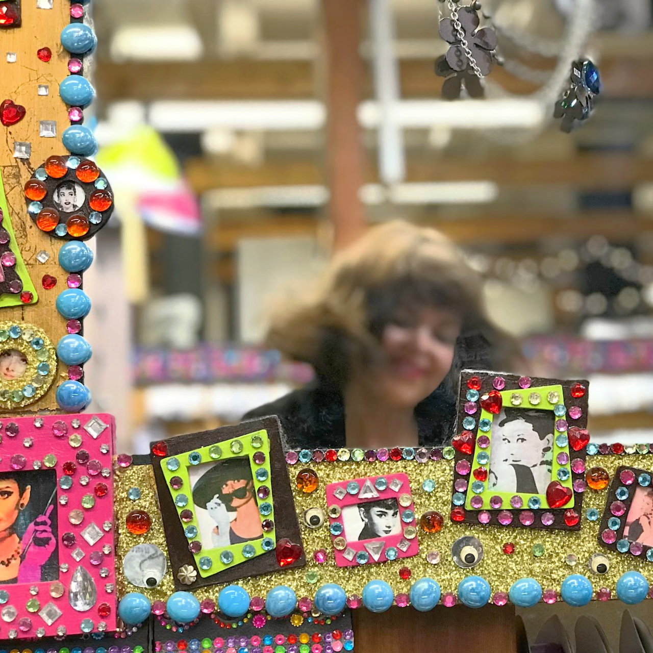 Colourful mirror frame with images of actress Audrey Hepburn surrounded by mini gem-embellished frames, with a blurry image of a smiling, dark haired woman.