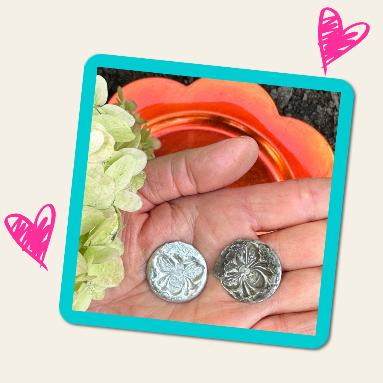 Photo of an open hand with two metal bee charms, with lime hydrangea blossoms on the left, and an orange plate and black dirt in the background.